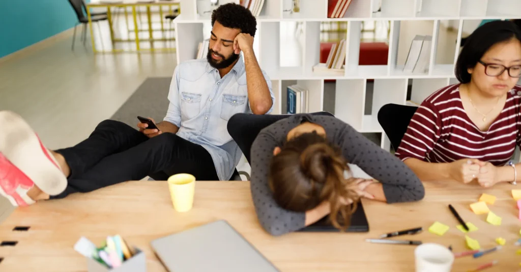 Three coworkers appearing burnt out in an office setting; one rests their head on the table, another is on the phone with a hand to their temple, and the third looks concerned while reviewing notes. White cubbies with books are in the background.